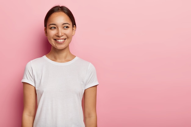  shot of happy young Asian woman has tender smile, looks aside with charming expression, wears casual white t shirt, has natural beauty, isolated on pink wall. People and emotions concept