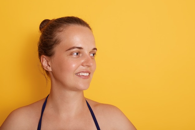 Shot of happy young Caucasian woman with tender smile, looks aside with charming expression, wears swimming suit, has natural beauty, isolated over yellow wall.