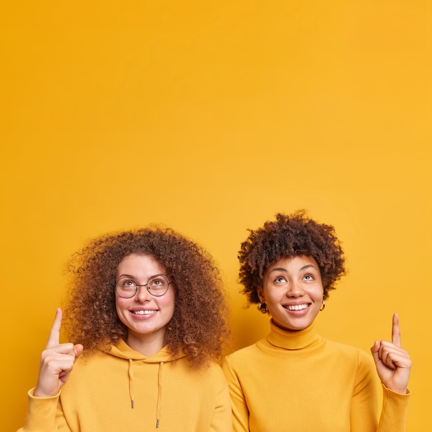 幸せな若い多様な女性のショットは、黄色の壁の上に隔離されたコピースペースの現在のアイテムまたは製品スタンドで上を指しています。人々のプロモーションと広告のコンセプト