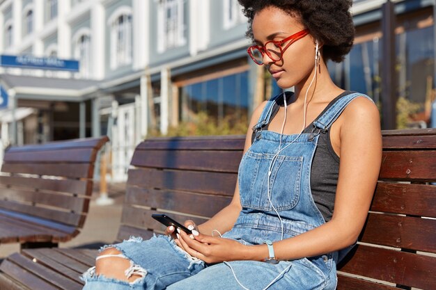 Colpo di ragazza hipster con pelle scura, taglio di capelli afro, chat con follower nei social network, ascolta la musica preferita in auricolari, trascorre il tempo libero all'aperto, si siede su una panca di legno, aspetta un amico