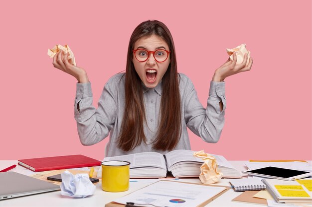 Shot of irritated young worker keeps crag of paper in hands, yells with depressed expression, has much work, reads books