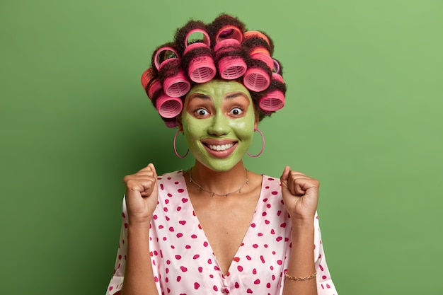 Shot of joyful adult woman anticipates something awesome happen, clenches fists with success, smiles broadly, applies moisturizing green mask for skin care, hair rollers on head, poses indoor