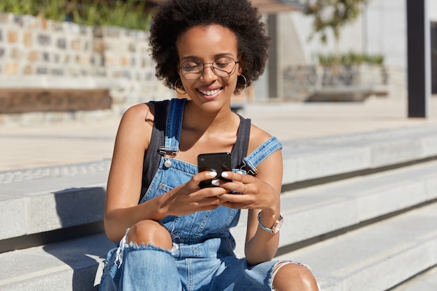 Shot of joyful teenager with dark skin, curly hair, reads comments on her blog, watches video online in social networks, wears casual denim dungarees, poses at stairs alone, connected to 3G.