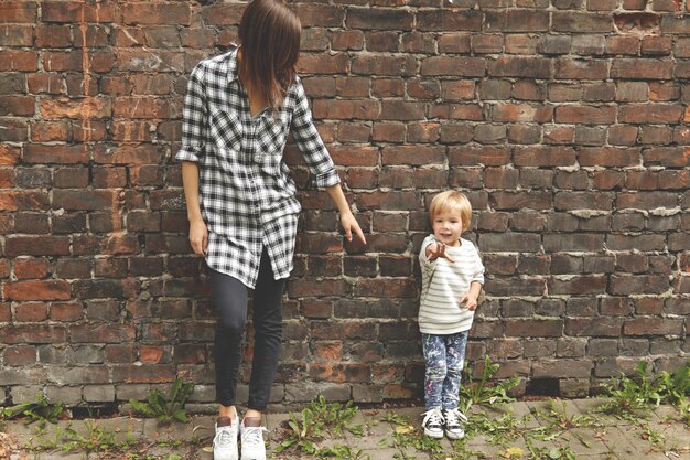 Shot of little boy with his caring sister near brick wall. Skinny Caucasian girl dressed in checked shirt, black trousers. She stretching arm to younger brother, but he pulling his hand ahead.