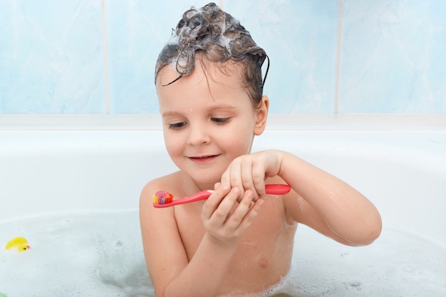 Shot of little child brushing her teeth while taking bath, charming wet lady holds red tooth brush