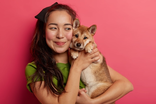 Shot of lovely korean girl being in love with her shiba inu dog, embraces pet with smile, has dark hair, wears green t shirt, poses with animal against pink background.