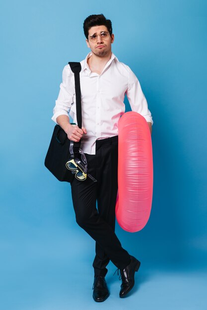 Shot of man in business suit, gathered on trip to sea. Guy in glasses poses with diving mask, inflatable mattress and bag with documents.