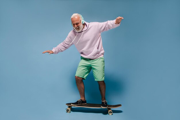 緑のショートパンツとスケートボードに乗ってパーカーの男のショットひげとヘッドフォンで孤立した背景にポーズをとってポジティブな男