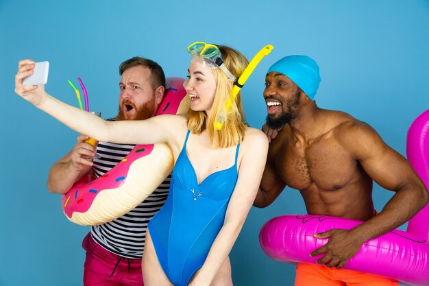 Shot for memories. Happy friends resting and take selfie or vlog on blue studio background. Concept of human emotions, facial expression, summer holidays or weekend. Chill, summertime, sea, ocean.