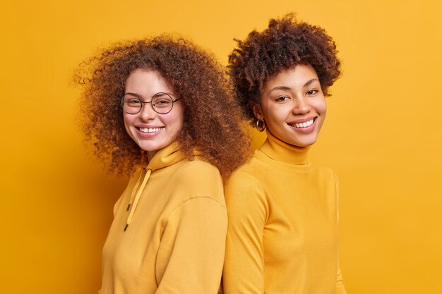  shot of mixed race women stand back to each other look with happy expressions feel satisfied wear casual clothes isolated over yellow wall. People emotions relationship concept.