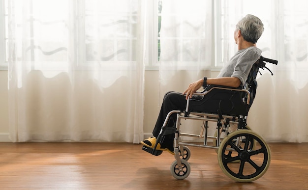 無料写真 車椅子に座っている年配の男性が車椅子で一人で家にいるホームエルダーのアジア人男性のショットは、リビングルームから窓の外の景色を見てください