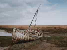 Бесплатное фото Снимок брошенной разбитой лодки, оставленной на берегу реки, в окружении пшеничного поля