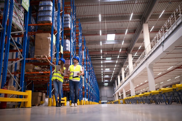Снимок двух рабочих, идущих по большому складскому центру, осматривающих стеллажи с товарами и планирующих доставку на рынок.