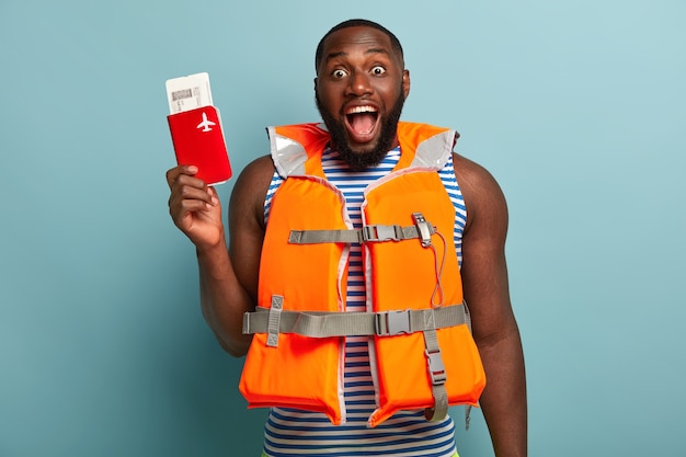 Shot of overjoyed dark skinned man with thick bristle, happy to have adventure journey soon, holds passport with tickets