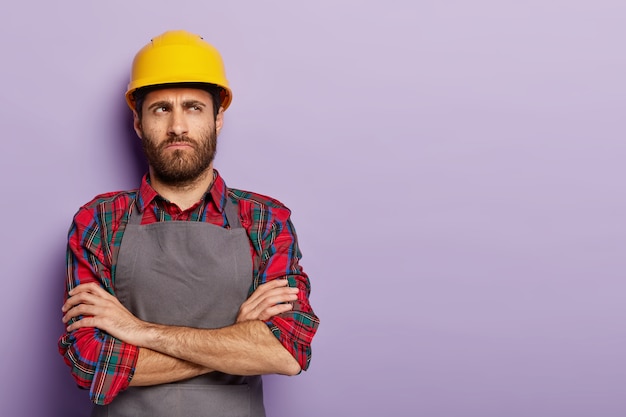 Free photo shot of pensive dissatisfied industrial worker wears yellow hardhat and apron