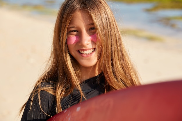 Colpo di donna soddisfatta dai capelli chiari con un sorriso a trentadue denti, ha zinco da surf sul viso per proteggersi dal sole, felice dopo il viaggio di surf con un amico