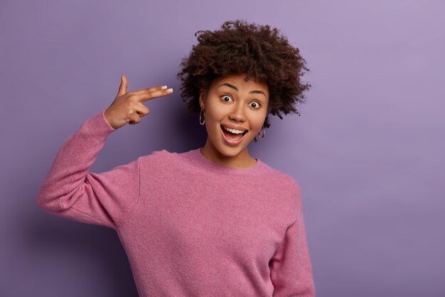 shot of pretty curly young African American woman foolishes around and makes finger gun pistol to temple, demonstrates suicide gesture, wears casual jumper, isolated over purple wall