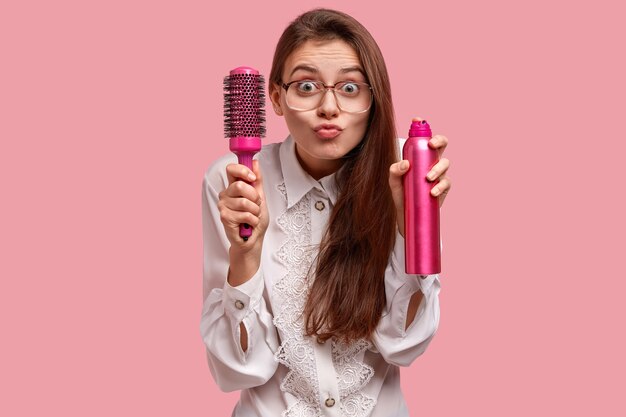 Shot of pretty young female model makes grimace, holds hairbrush and spray, has curious expression, going to make hairstyle