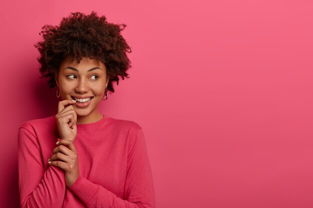 アフロの髪のかなり若い女性のショット、人差し指を口の近くに保ち、脇に集中し、幸せな表情を持ち、何か魅力的なものに気づき、ピンクの壁に隔離され、空きスペース