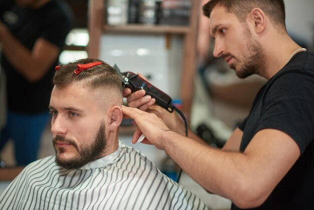 仕事でプロの床屋のショット。ハンサムな若い男が地元の理髪店で散髪を取得します。