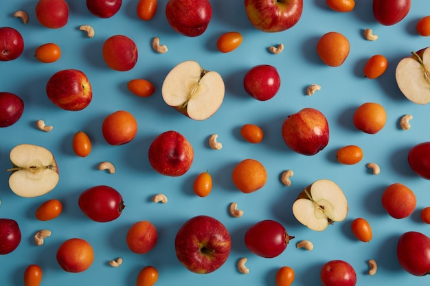 Выше выстрел из красных спелых яблок, персиков, томарилло, кумквата и питательных орехов кешью на синем фоне. Креативная композиция из вкусных фруктов. Сладкая еда с виаминами, концепция здорового питания