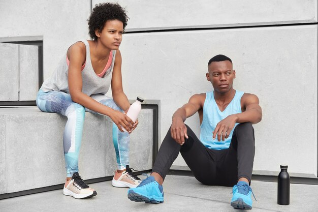 Shot of self confident black woman and man dressed casually, sit at stairs near wall, drink fresh water not to feel thirsty, have rest, lead healthy lifestyle, have exercises outside. Horizontal view