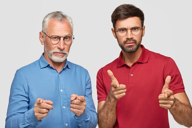 снимок серьезных уверенных в себе партнеров-мужчин разного возраста, которые указывают прямо, делают выбор, носят строгую синюю рубашку и красную яркую футболку, позируют вместе у белой стены.