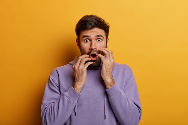  shot of shocked scared man being in panic, keeps hands near opened mouth, sees something terrible, feels excited, wears purple sweatshirt, isolated on yellow wall, hears awful news