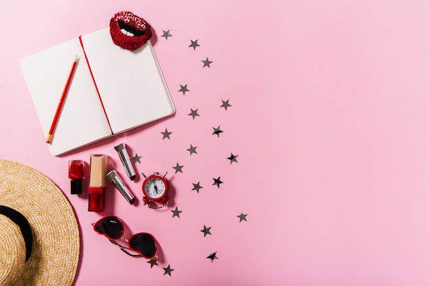 Shot of straw wide-brimmed hat, set of cosmetics, sunglasses and notebook on pink wall,