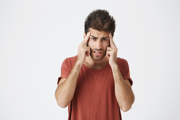  shot of stylish bearded Caucasian man holding fingers on his temples with suffering expression, as if trying to remember something important or having headache