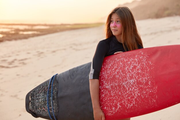 Shot of surfer woman in black swimsuit, holds surfboard, has stroll across sea or ocean