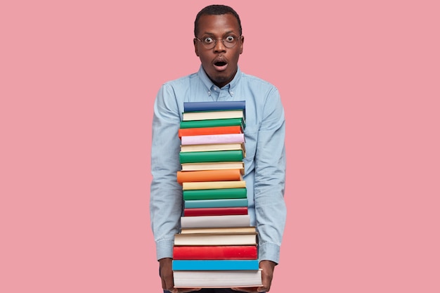 Colpo di sorpreso uomo afroamericano trasporta una pila di libri colorati, spaventato di avere una scadenza, vestito con abiti formali, mantiene la bocca aperta