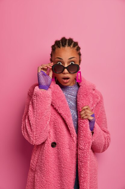  shot of surprised Afro American woman looks from sunglasses with shocked expression, cannot believe her eyes, dresses winter coat and sport gloves, stunned by big discounts in shopping mall