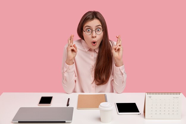 shot of surprised european woman with astonished facial expression, keeps hands in praying gesture, dressed in elegant shirt, being office pefectionist