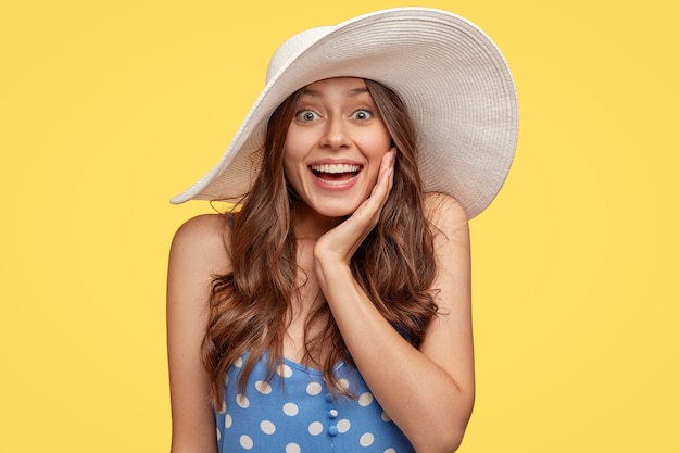Shot of thrilled smiling young lady has natural dark hair, white teeth, broad smile, touches cheek with hand, wears stylish summer hat, feels amazed by great news, isolated over yellow wall