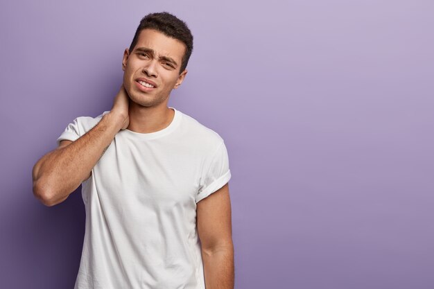 shot of tired overworked young man needs massage, feels pain in neck, sitting hardworking long time, wears t shirt, expresses negative emotions, isolated over purple wall, free space.