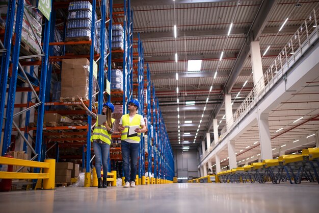 Shot of two workers walking through large warehouse center, observing racks with goods and planing distribution to the market