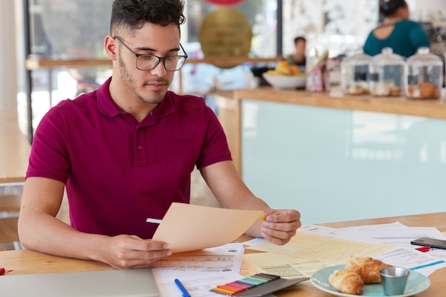 Shot of unshaven guy examines paperwork, uses stickers, dressed in casual t shirt and spectacles. Creative male blogger works with documentation, has hard working day, develops new strategy.