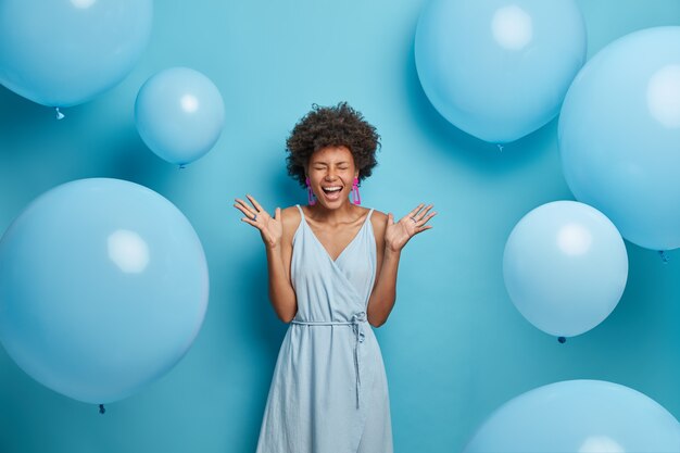 明るい陽気な暗い肌の女性のショットは非常に幸せで興奮していると感じ、手のひらを上げて笑い、パーティーで自由な時間を過ごし、イヤリングとリングが付いた素敵な青いサマードレスを着て、風船の近くでポーズをとる