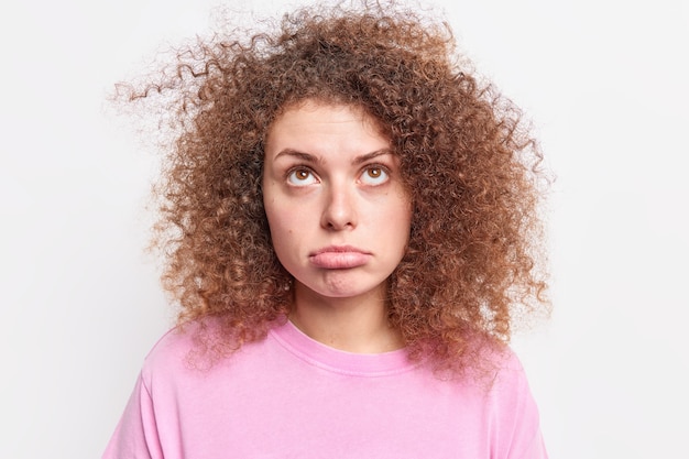 Free photo shot of upset european woman purses lower lip looks sadly above being disappointed by something has curly bushy hair dressed casually isolated over white wall. face expressions.