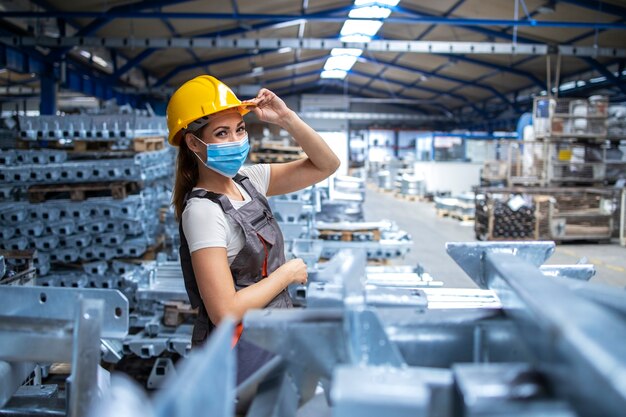 Shot of woman factory worker in uniform and hardhat wearing face mask in industrial production plant