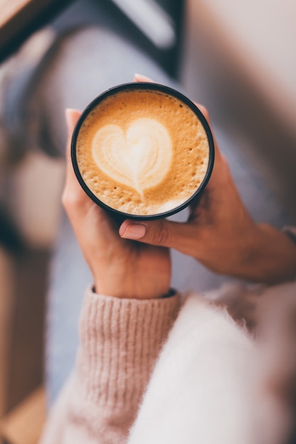 Shot of woman hands hold cup of hot coffee with heart design made of foam.