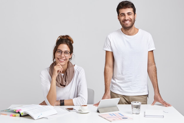 shot of woman and man coworkers in white clothes, look with glad expressions, work on common task together, pose near workspace, isolated