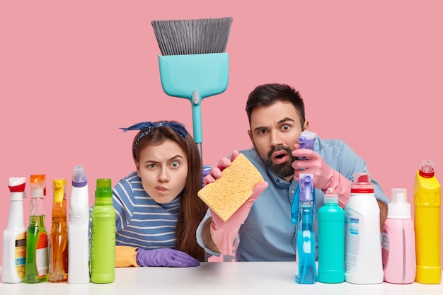 shot of woman and man look scrupulously, do housework, clean everything, hold sponge and broom, sit at workplace with detergents, isolated over pink wall. Household duties