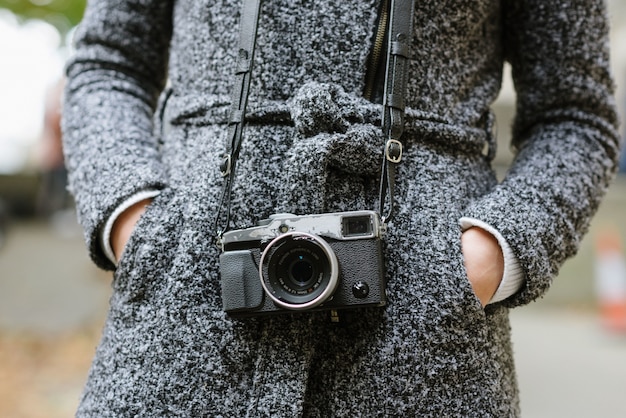 Shot of a woman standing with her hands in her pockets wearing a gray coat and a vintage camera