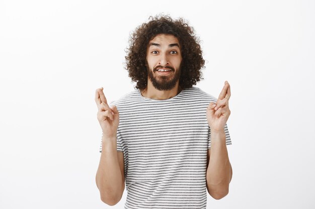 Shot of worried handsome bearded Eastern male model with afro hairstyle in striped t-shirt, raising crossed fingers and hoping, making wish and begging faith to fulfill it