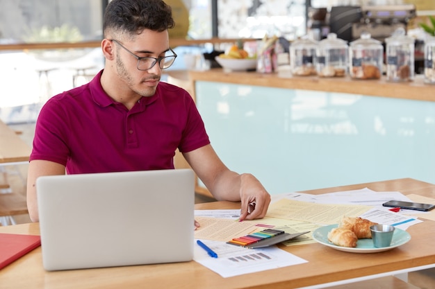 Shot of young male uses laptop computer for checking exchange rate in internet, works with financial documents, leaves stickers on some of papers, going to have tasty lunch in cozy snack bar