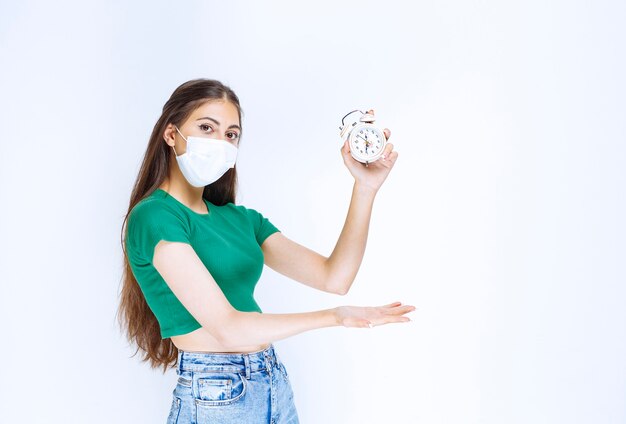 Shot of young woman in medical mask showing alarm clock.