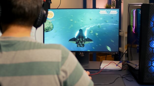 Over shoulder footage of professional streamer playing digital space shooter video games on computer using headphones, microphone and mouse