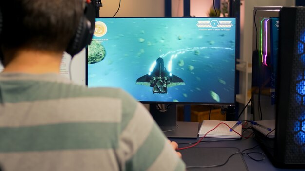 ヘッドフォン、マイク、マウスを使用してコンピューターでデジタルスペースシュータービデオゲームをプレイするプロのストリーマーの肩越しの映像